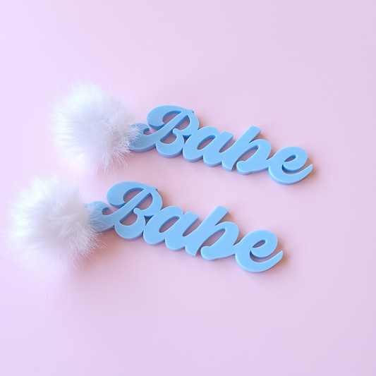 Two blue 'Babe' earrings with fluffy white pom poms on a pink background
