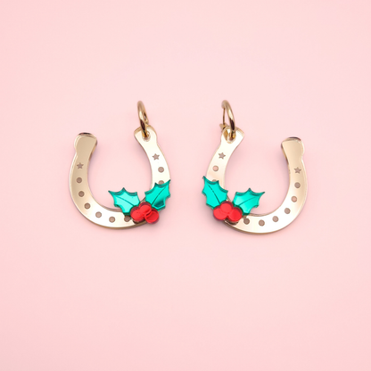 Gold acrylic horseshoe Christmas earrings with green holly leaves and red berries on a pink background.