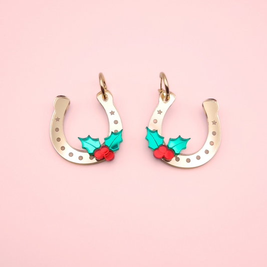 Gold acrylic horseshoe Christmas earrings with green holly leaves and red berries on a pink background.