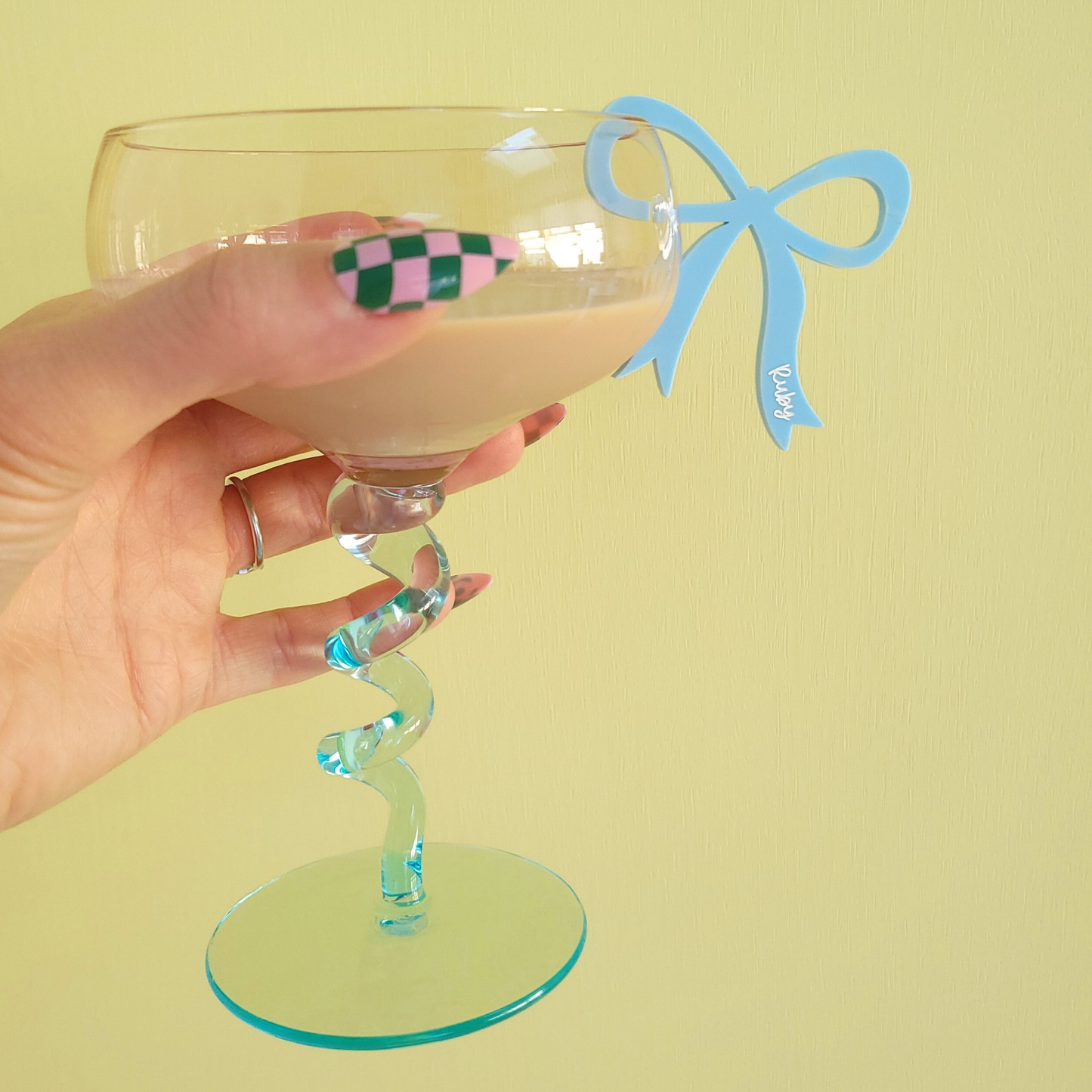 Hand holding a glass with a blue acrylic bow drink charms with personalised name Ruby on yellow background