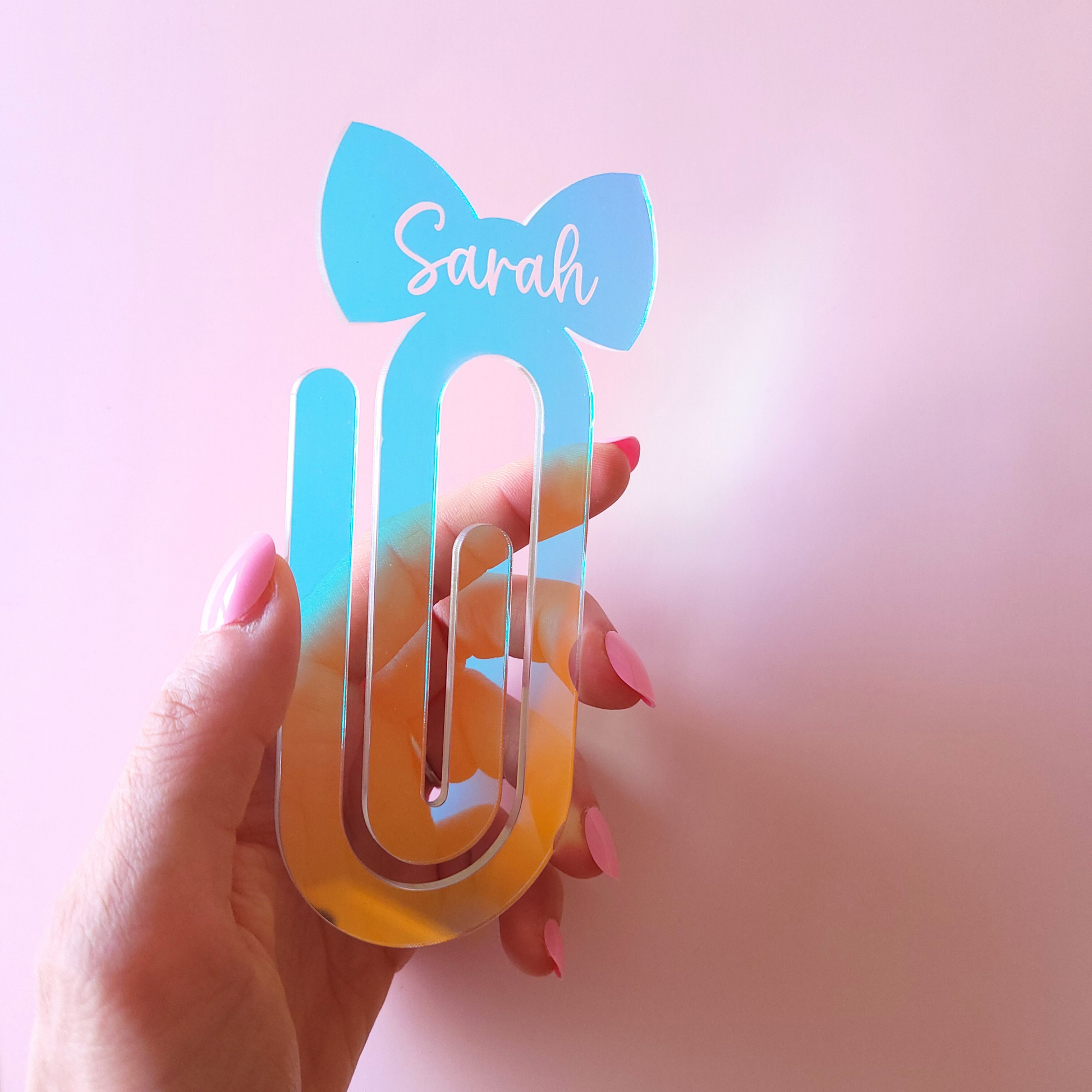 Iridescent girly acrylic bow bookmark with custom name Sarah held by a hand. 