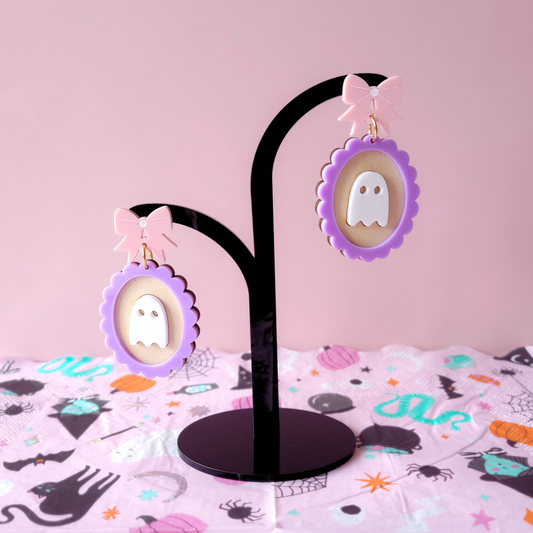 A pair of pastel ghost earrings with white ghost motifs inside lavender scalloped frames, topped with pink bow studs, displayed on a Halloween-themed backdrop.