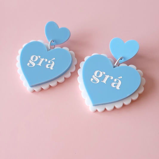Pair of pastel blue acrylic heart earrings with “grá” text on pink background