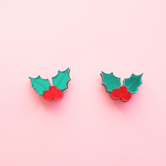 Small acrylic holly berry stud earrings with red berries and green leaves on a pink background