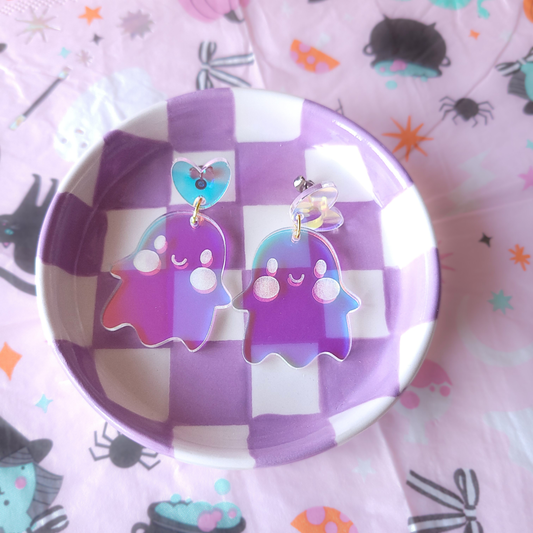 Pair of holographic acrylic ghost earrings with smiling faces, dangling from colorful heart studs, displayed in a checkered jewellery tray.