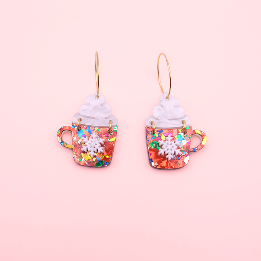 Rainbow acrylic hot cocoa mug earrings with whipped cream tops and snowflake detail on a pink background.