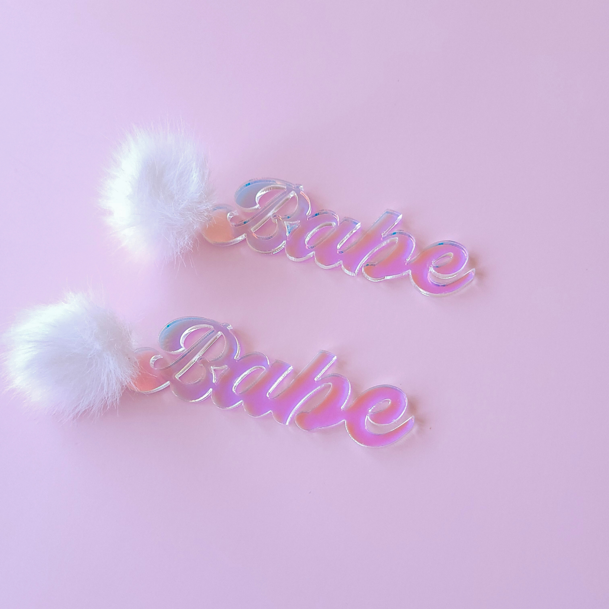 Iridescent 'Babe' earrings with white fluffy pom-poms on a pink background