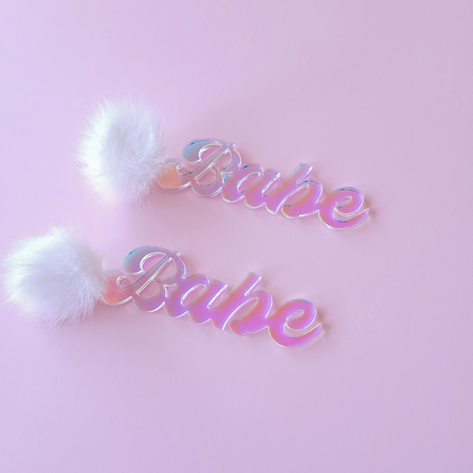 Iridescent 'Babe' earrings with white fluffy pom-poms on a pink background