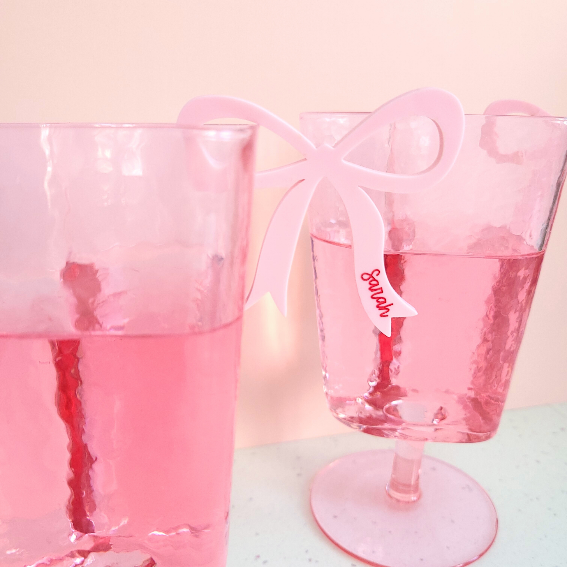 Close up of a pink acrylic bow drink charm with personalised Sarah clipped on a pink glass