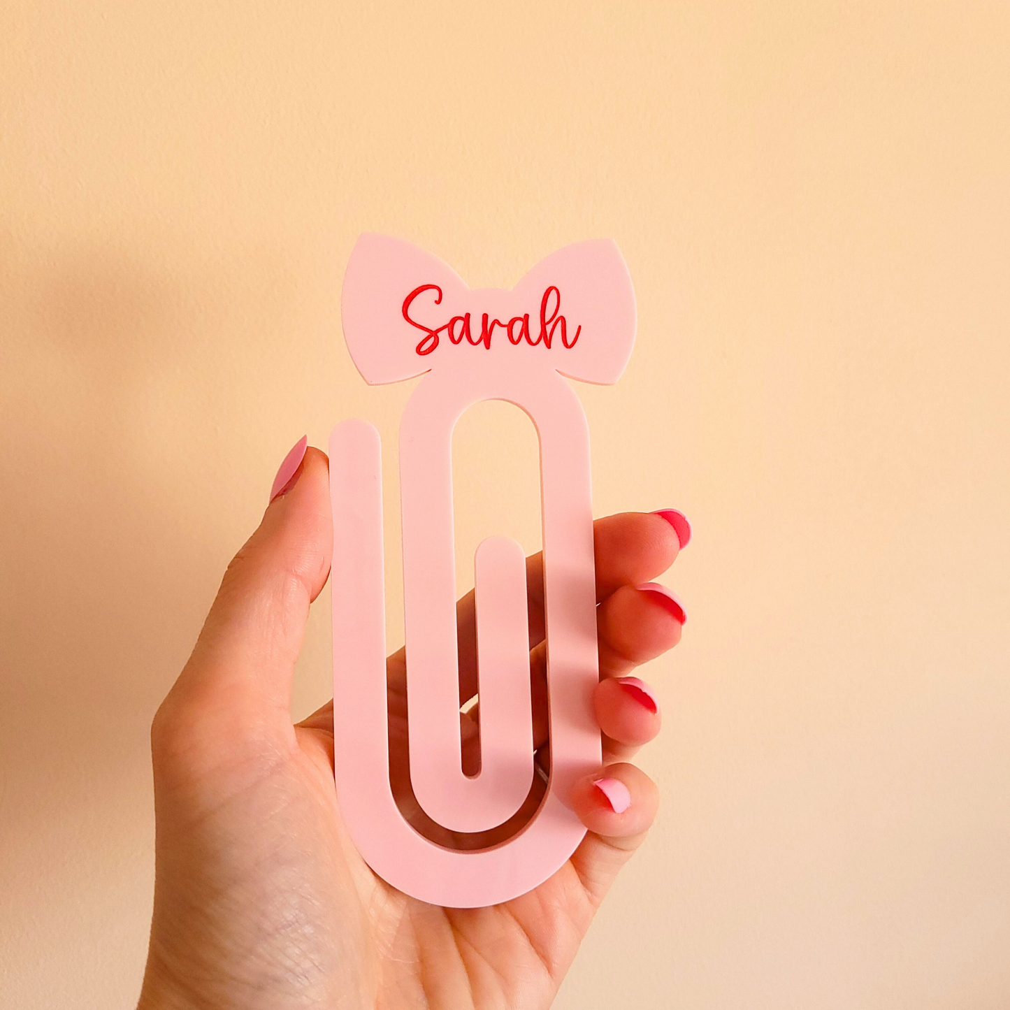 Hand holding pink acrylic bow bookmark with custom name Sarah in red text.