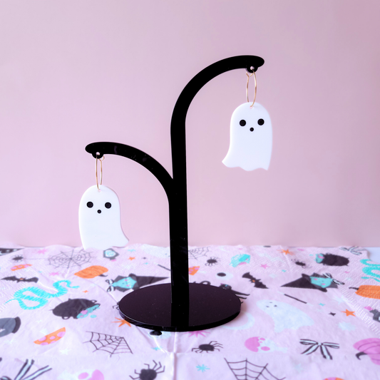 A pair of white acrylic ghost earrings with black eyes and a gold hoop, displayed against a pink background with Halloween-themed fabric