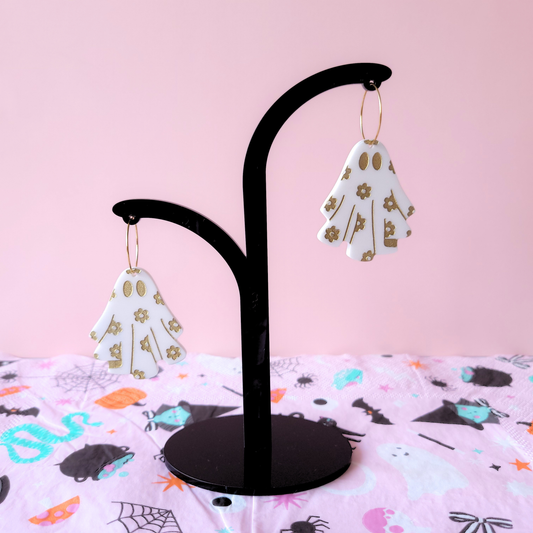 Pair of white acrylic ghost earrings with gold flower details, displayed on a black earring holder on a pink background.