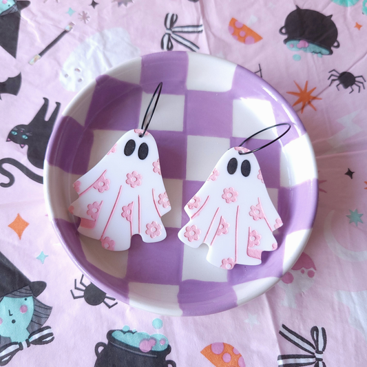 Pair of white acrylic ghost earrings with pink flower details, displayed in a purple checkered dish on Halloween-themed fabric background.