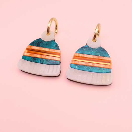 Teal marbled acrylic winter hat earrings with orange mirror stripes and beige glitter trim on gold hoops — handmade festive jewellery.