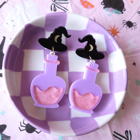 Pair of purple acrylic potion bottle earrings with red glitter potion and black witch hat studs, displayed in a purple checkered dish on Halloween-themed fabric.